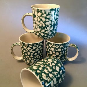 May include: Four ceramic mugs with a cream-colored interior and handles. The exterior features a white base with a speckled green pattern. The mugs are stacked, with one tilted on its side, showcasing the design and shape.
