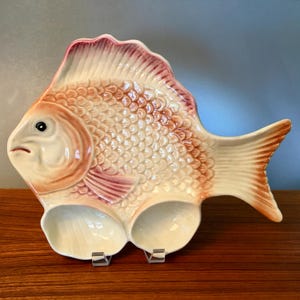 May include: A ceramic fish-shaped serving dish with a cream-colored body, orange scales, and pink accents. The dish has two circular compartments and a detailed fin and tail design. The dish is displayed on a wooden surface.