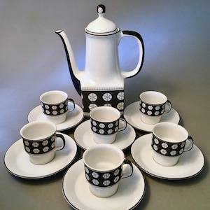 May include: A white and black ceramic coffee set. The set includes a tall coffee pot, six cups, and six saucers. The cups and the coffee pot have a black band with a white circular pattern. The saucers have a black trim.