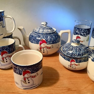 May include: A collection of winter-themed ceramic tableware featuring a snowman design. The set includes mugs, a teapot, sugar bowl, creamer, and glasses. The pieces are white with blue speckled detailing and a snowman illustration wearing a red hat and scarf.
