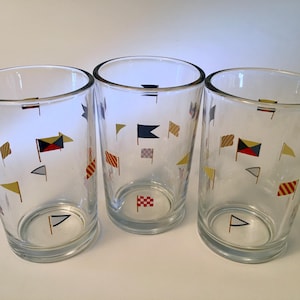 May include: Three clear glass tumblers featuring a repeating pattern of colorful nautical signal flags. Each cylindrical drinking glass displays various flag designs in red, blue, yellow, and white, arranged in a scattered layout around the exterior.