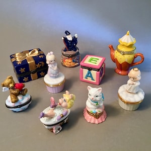 May include: A collection of decorative trinket boxes in various shapes and designs. Includes a blue gift box, a wizard, a teapot, a teddy bear holding a heart, a baby block, a cat, and figures of children. The boxes are made of porcelain and have gold accents.