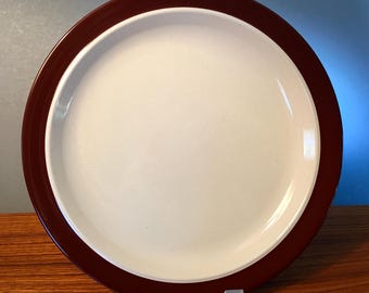 May include: A round, ceramic plate with a cream-colored center and a dark brown rim. The plate is displayed on a clear plastic stand, showcasing its simple, yet elegant design. This plate is suitable for serving meals.