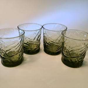 May include: A set of four short drinking glasses made of translucent olive green glass. Each glass features a textured, wavy, and organic surface pattern that creates a rippled effect across the exterior, with a clear rim and a solid base.