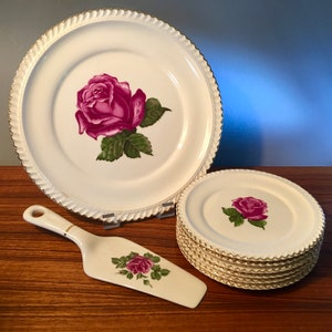 May include: A vintage white porcelain dinnerware set featuring a large plate, several smaller plates, and a cake server. Each piece is adorned with a vibrant magenta rose and green leaves. The edges have a gold trim and a textured design.