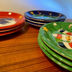 May include: A stack of colorful ceramic plates with hand-painted designs. The plates are red, blue, and green, each featuring unique illustrations of stars and other festive elements. The plates are stacked on a wooden surface.