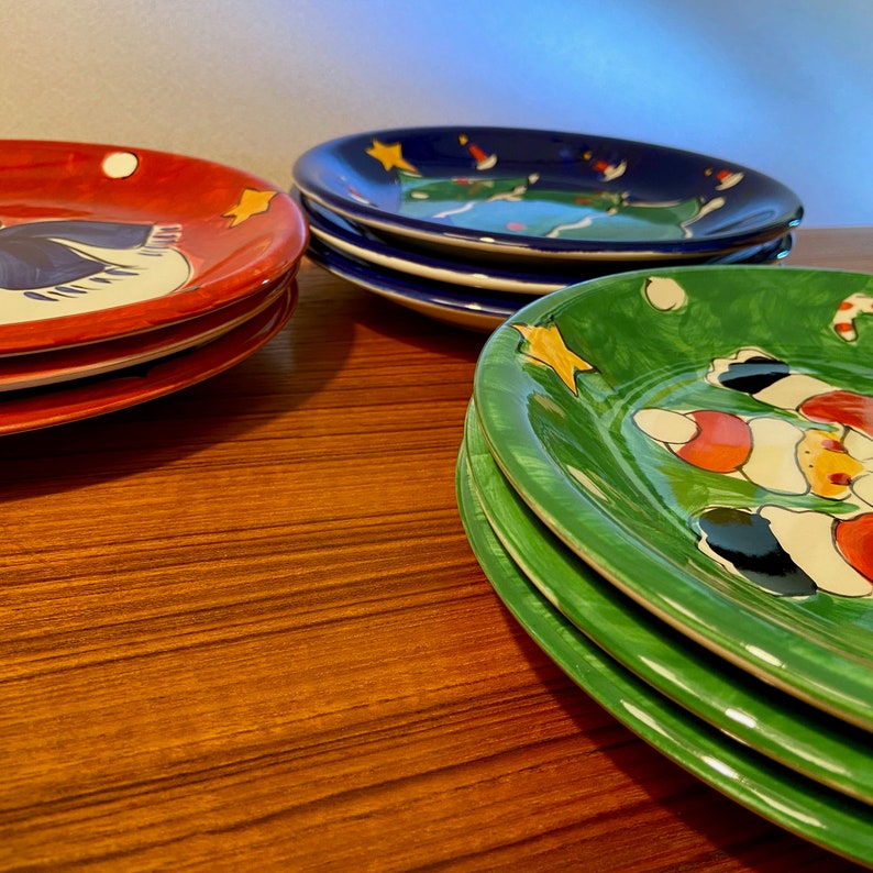 May include: A stack of colorful ceramic plates with hand-painted designs. The plates are red, blue, and green, each featuring unique illustrations of stars and other festive elements. The plates are stacked on a wooden surface.