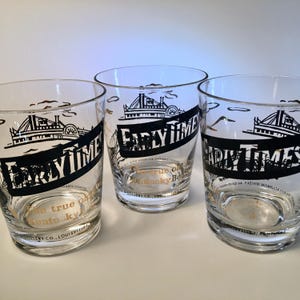 May include: Three clear drinking glasses with a black and gold design. The glasses feature the text "Early Times" in a black banner, along with an illustration of a steamboat and bridge. The bottom of the glasses have gold text.