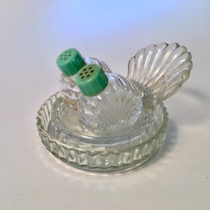 May include: A clear glass salt and pepper shaker set shaped like a turkey, featuring two light green plastic screw-on lids with small holes. The shakers rest on a matching circular glass base with a ribbed, scalloped edge design.