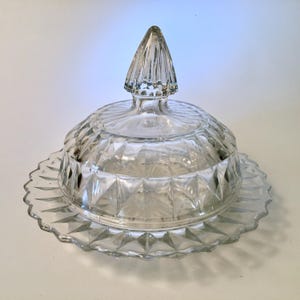 May include: A clear pressed glass butter dish featuring a dome-shaped lid with a pointed finial handle. The lid and the circular base plate are decorated with a repeating diamond pattern and a scalloped edge, creating a textured, reflective surface.