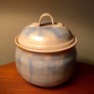 May include: A handmade ceramic jar with a lid. The jar is a light beige color with blue brushstrokes and a matching lid. The lid has a curved handle and decorative accents. The jar is sitting on a wooden surface.