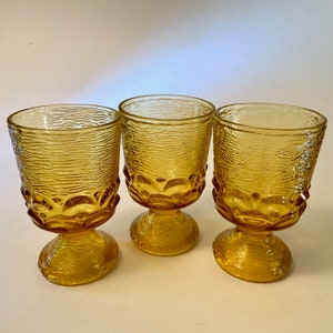 May include: Three amber-colored, textured glass goblets. Each goblet has a textured, cylindrical bowl and a rounded base. The goblets are made of a transparent, golden-yellow glass.