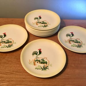 May include: A stack of cream-colored plates with a yellow rim and a rooster design. The rooster is red and green, standing on a brown basket with green foliage. Several plates are stacked, with others scattered around.
