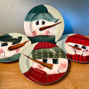 May include: Four ceramic snowman plates. Each plate features a snowman face with a carrot nose, black eyes, and a red or green scarf and hat. The plates have a light gray background with white polka dots.