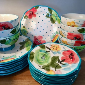 May include: A collection of white ceramic dinnerware with a floral design. The plates and bowls feature a turquoise rim and polka dots. The floral pattern includes red, green, and yellow flowers.