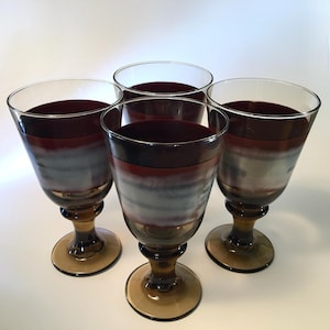 May include: Four vintage wine glasses with a unique design. The glasses feature a layered effect with a dark red top, a gray middle section, and a clear base. The stems and bases are a warm amber color, creating an elegant aesthetic.