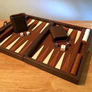 May include: A brown leather backgammon set with a felt playing surface. The board features alternating brown, tan, and white triangles. Includes dice, dice cups, and game pieces. The set is open and ready for play.