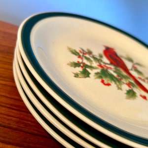 May include: A stack of white ceramic plates with a green border. Each plate features a red cardinal bird perched on a holly branch with red berries. The plates are stacked on a wooden surface.