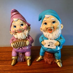 May include: Two ceramic garden gnomes. One gnome is dressed in pink and purple, playing an accordion. The other gnome is in blue, singing from a sheet of music. Both gnomes have white beards and gold boots.