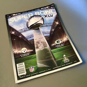 May include: A Super Bowl XLV game program featuring the Vince Lombardi Trophy centered against a stadium background. The cover displays the Pittsburgh Steelers and Green Bay Packers logos, the NFL shield, and the text North Texas, Sunday, February 6, 2011.