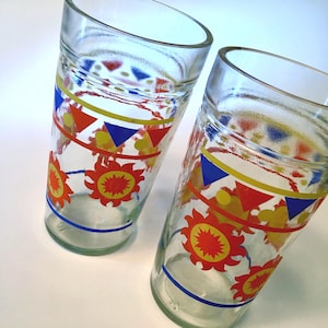 May include: Two clear glass tumblers featuring a colorful printed design. The pattern includes a horizontal yellow line, blue and red triangles, yellow circles, and orange sun-like shapes with red centers, finished with a thin blue line at the base.