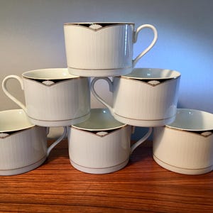May include: A stack of six white teacups with a black and gold geometric design. The cups have a vertical ribbed texture and a gold trim. The handles are curved. The cups are stacked on a wooden surface.