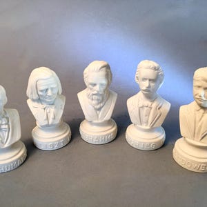 May include: Five white ceramic busts of composers, including Schumann, Liszt, Brahms, Grieg, and McDowell. Each bust is on a pedestal with the composer's name. The busts are detailed and have a classical aesthetic.