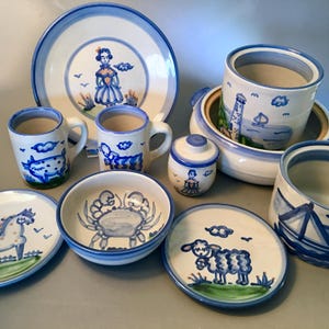 May include: A collection of hand-painted ceramic tableware with a nautical theme. The set includes plates, bowls, mugs, and a lidded jar, all featuring blue and white designs of a crab, sheep, lighthouse, and figures. The rims and edges are accented with blue.