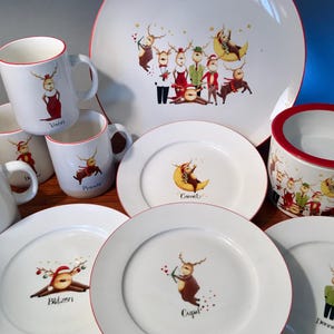 May include: A collection of white ceramic tableware with a festive reindeer theme. Includes mugs, plates, and a bowl, all with red trim. Each piece features a different reindeer character illustration, perfect for holiday gatherings.