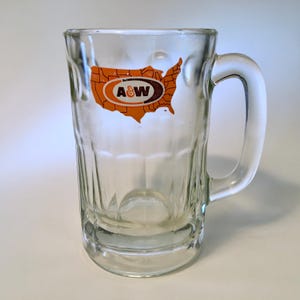 May include: A clear glass beer mug featuring a thick handle and a vertical paneled design. The front of the mug displays an orange graphic of the United States map with the brown and white A&W logo centered within the map outline.