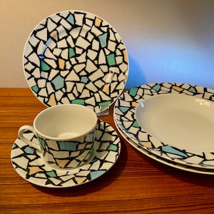 May include: A white ceramic dish set with a mosaic pattern in black, light blue, green, and tan. The set includes a dinner plate, a saucer, a teacup, and a serving dish. The dishes are arranged on a wooden surface.