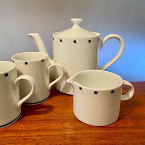 May include: A white ceramic tea set with black square accents. The set includes a teapot with a lid, two mugs, and a creamer. The mugs and creamer have a black stripe near the base. The set is on a wooden surface.