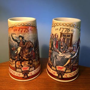 May include: Two ceramic beer steins with historical illustrations. One depicts a man on horseback, the other a group of men around a table. Both steins have the text "1775" and "1776" and the words "Birth of a Nation".