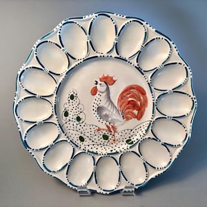 May include: A white ceramic egg serving platter featuring a central hand-painted illustration of a rooster with orange and black details. The plate has twelve oval indentations around the perimeter, each outlined with a dark teal painted border.