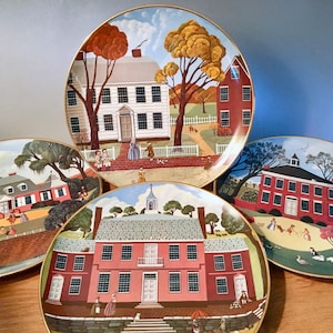 May include: Four decorative plates with gold rims, each featuring a detailed illustration of a colonial-era building and landscape. The plates showcase a variety of architectural styles, with figures and trees adding depth to the scenes. The color palette includes reds, whites, and browns.