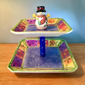 May include: A two-tiered serving tray with a snowman figurine. The tray has a blue center with white polka dots and a colorful border. The snowman has a black top hat, a red scarf, and yellow arms. The stand is blue.
