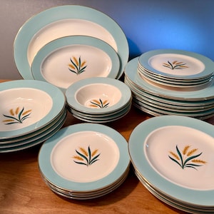 May include: A collection of vintage dinnerware with a light blue border and gold trim. Each piece features a wheat stalk design in the center. Includes plates, bowls, and serving dishes.