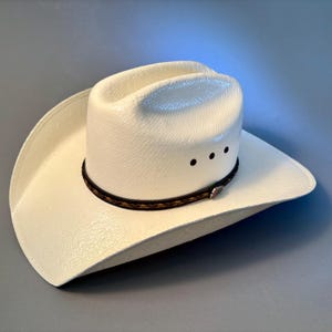 May include: A white cowboy hat with a textured finish, featuring a cattleman crease crown and a wide, curved brim. The hat is accented with a dark brown braided leather hatband that includes a small silver-toned metal detail on the side.