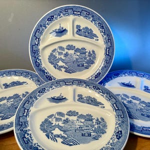 May include: A set of four white divided plates with a blue Willow pattern. Each plate features three compartments and a detailed landscape design with a bridge, boat, and pagoda. The plates have a scalloped edge.