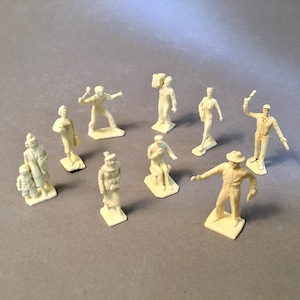 May include: A collection of small, off-white plastic figurines depicting various figures in different poses. The figurines include people carrying objects, gesturing, and wearing hats. The figures are on individual rectangular bases.
