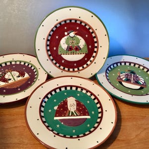 May include: A set of four decorative ceramic plates with a winter holiday theme. Each plate features a snowman or winter scene in the center, surrounded by a red, green, or purple border with white stars and black dots. The plates have a cream-colored rim.
