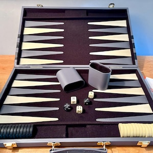May include: Open backgammon set with a gray wooden case, featuring a dark purple playing surface. The game pieces are black and cream-colored. Dice and a container are also included. A booklet with the text "Backgammon Rules" is visible.