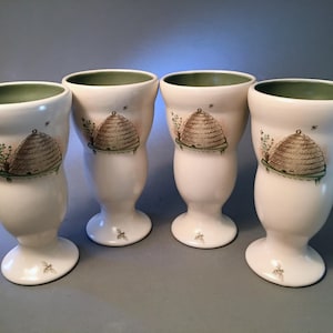 May include: Four white ceramic goblets with a green rim and a beehive design. Each goblet has a beehive illustration with brown and green details, along with small bee and floral accents. The goblets have a pedestal base.