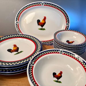May include: A white ceramic dinnerware set featuring a rooster design. The set includes an oval platter, plates, and bowls. Each piece has a red, green, and blue checkered border. The rooster is depicted in shades of red, brown, and black.