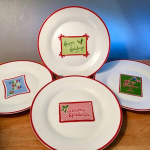 May include: Four white ceramic plates with red rims. Each plate features a holiday greeting in a colorful rectangular frame. The phrases include "Happy Holidays," "Happy New Year," "Merry Christmas to All," and "Season's Greetings."