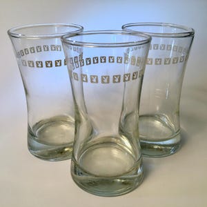 May include: Three clear glass tumblers with a flared shape. Each glass features a band of repeating gold-colored bunny logos near the top. The glasses are empty and appear to be vintage.