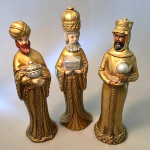 May include: Three gold-colored Wise Men figurines. Each figure is dressed in ornate robes and headwear, holding gifts. The figurines have detailed facial features and are painted with gold, red, and silver accents. A religious Christmas decoration.