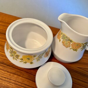K&ouml;nnte beinhalten: Wei&szlig;er Keramik-Zuckertopf mit Deckel und Sahnek&auml;nnchen. Beide Teile sind mit einem Blumenmuster in Gelb, Gr&uuml;n und Orange verziert. Der Zuckertopf ist offen, und das Sahnek&auml;nnchen hat einen Griff.