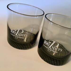May include: Two gray whiskey glasses with a silver rim. The glasses have a textured base and the text "100 Pipers Scotch by Seagram" printed in white. The glasses are angled.