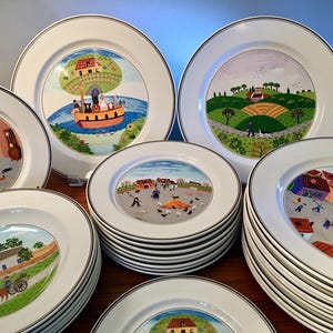 May include: A collection of white ceramic plates with black trim, each featuring a colorful, whimsical illustration. The plates depict various rural scenes, including a hot air balloon, a village, and a horse-drawn cart. The plates are stacked and arranged.